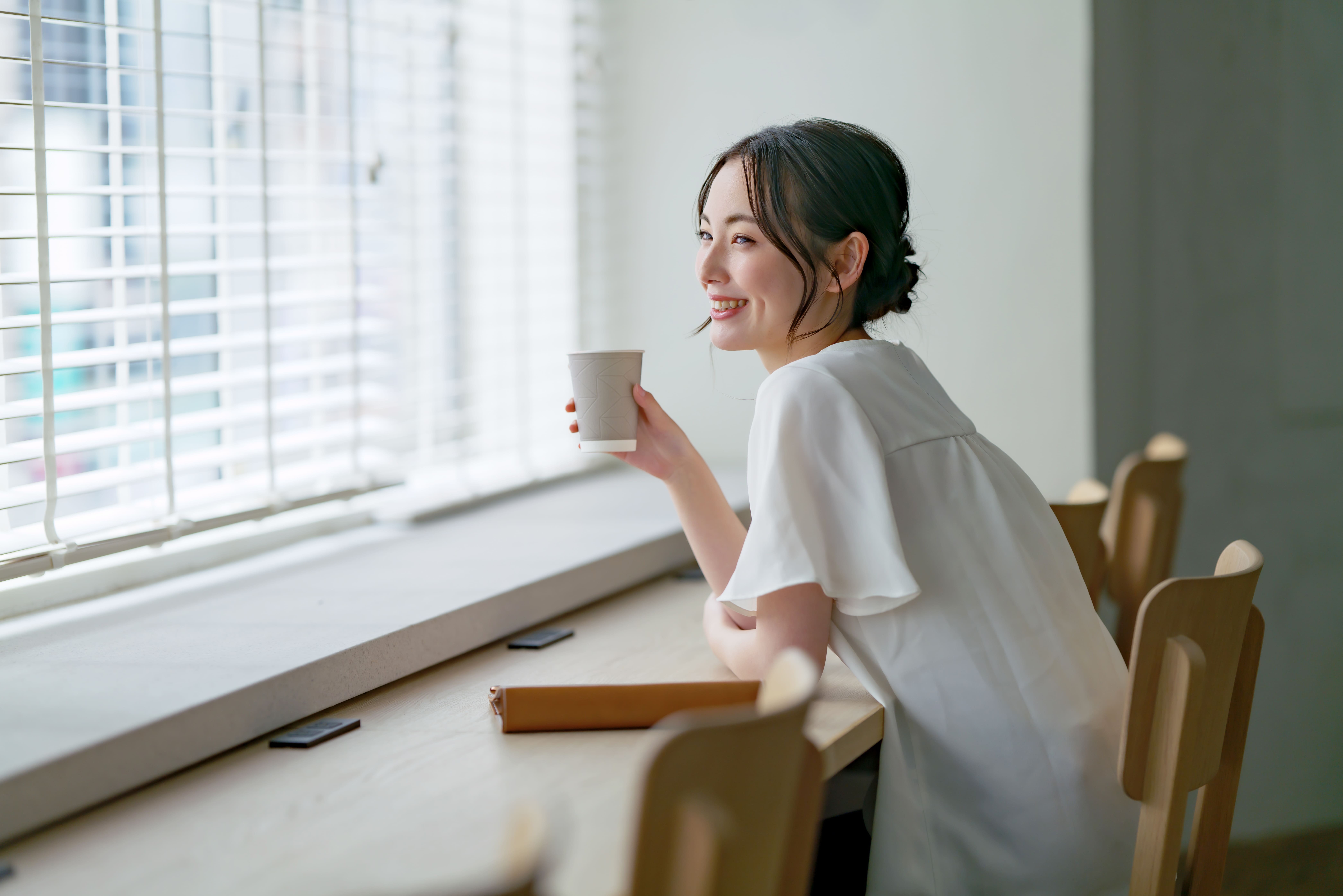 オフィスのカフェスペースで窓際に座り、紙カップのコーヒーを楽しむビジネスパーソン