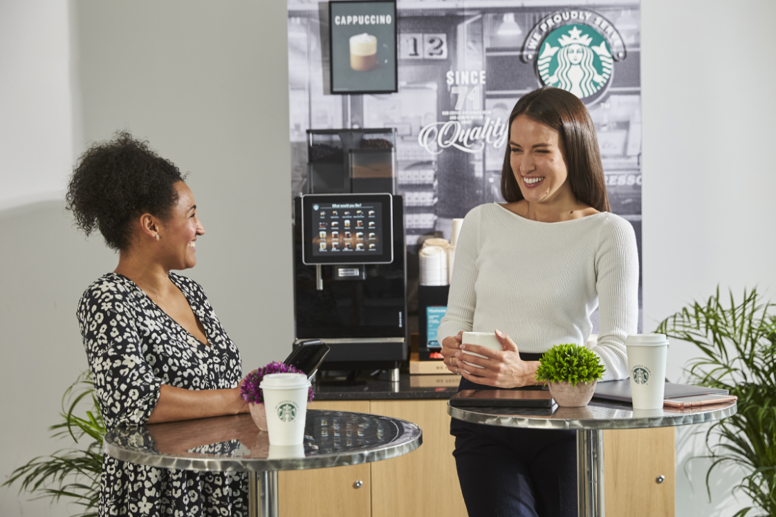 We Proudly Serve Starbucks® のコーヒーマシンとポスターの前でロゴが入った紙カップを持っている2人の人物