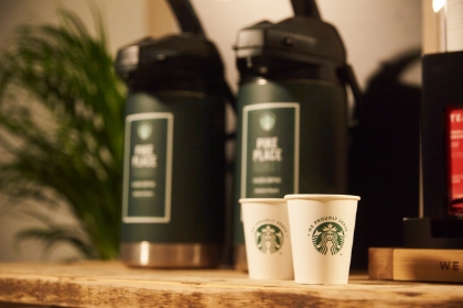 テーブルに置かれた２つのWe Proudly Serve Starbucks®の紙カップと背景にある専用の大きな2本のコーヒーポット